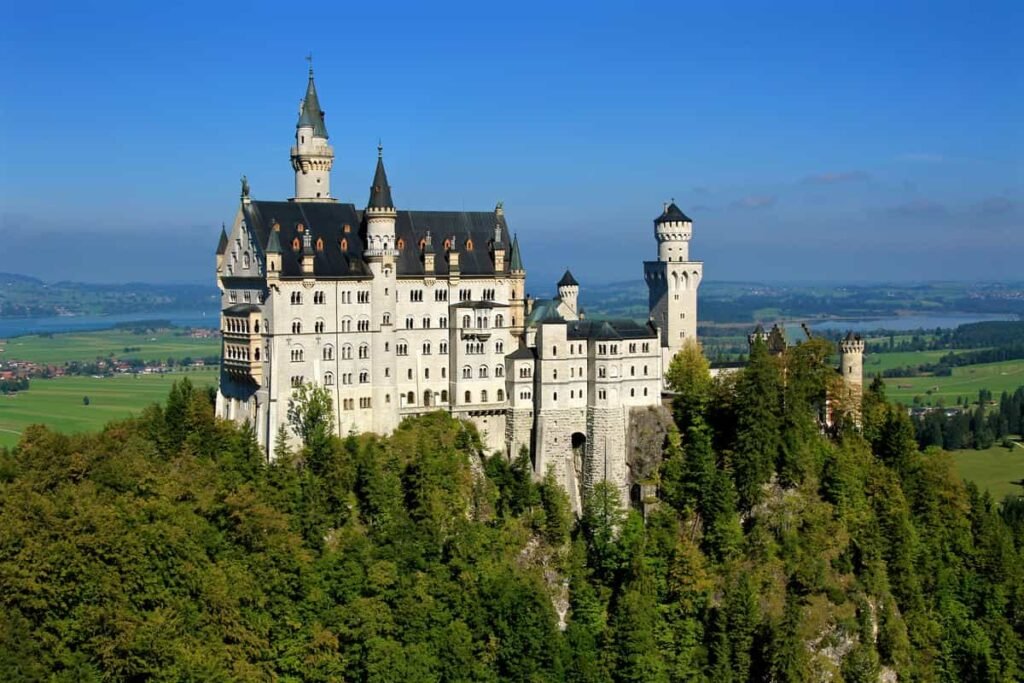 Neuschwanstein Castle perched on a forested hill in Bavaria, Germany, overlooking green fields and blue sky.