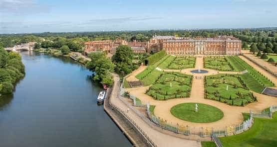 An aerial view of Hampton Court Palace with its detailed brick facade, manicured gardens, and a river flowing beside it. The surrounding area is covered with lush green trees.