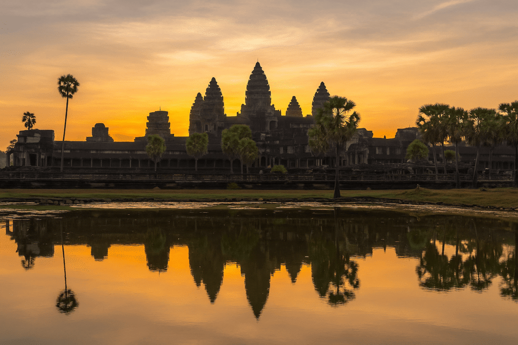 Sunrise view of Angkor Wat with temple towers reflected in the water and palm trees in the foreground.