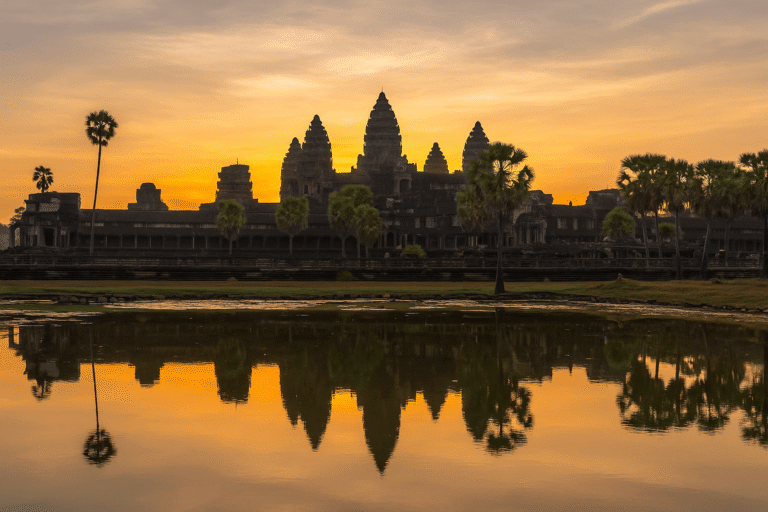 Sunrise view of Angkor Wat with temple towers reflected in the water and palm trees in the foreground.